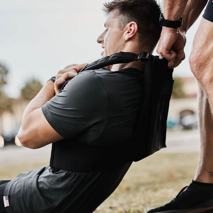 A man in exercise clothing is performing a sit-up outdoors with the help of another person holding a strap attached to him. He’s wearing the GORUCK Training Weight Vest 2.0 for added resistance. They are on a grassy area, and the assisting person is partially visible, focusing on their hands.