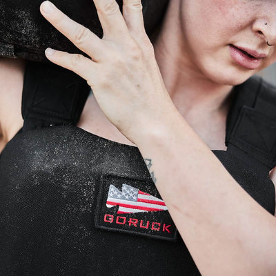 A person wearing the GORUCK Training Weight Vest 2.0, adorned with the American flag and the GORUCK logo, carries a black sandbag on their shoulder. Dust clings to the vest, while a small tattoo adorns their arm, highlighting their determination.