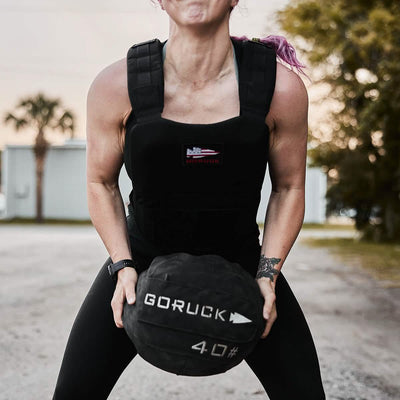 A person outfitted in a GORUCK Training Weight Vest 2.0 holds a 40-pound GORUCK exercise ball while standing on a gravel path, surrounded by trees and buildings outdoors.