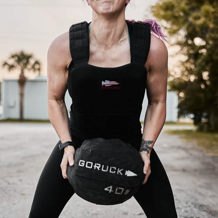 A person outfitted in a GORUCK Training Weight Vest 2.0 holds a 40-pound GORUCK exercise ball while standing on a gravel path, surrounded by trees and buildings outdoors.
