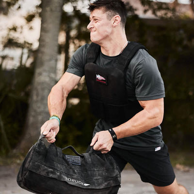 A man sporting the GORUCK Training Weight Vest 2.0 is outdoors, engaging in a fitness routine with a heavy bag. Clad in a dark athletic outfit, he looks concentrated among the trees and a softly blurred backdrop.