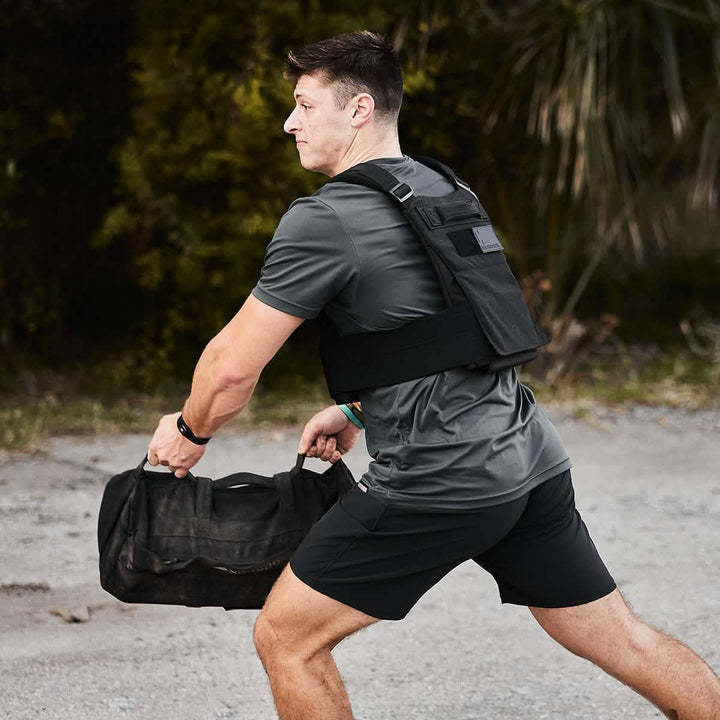 A man in athletic gear exercises outdoors, wearing the GORUCK Training Weight Vest 2.0. He holds a black duffel bag with one hand and appears to be mid-action, set against a natural backdrop of greenery.