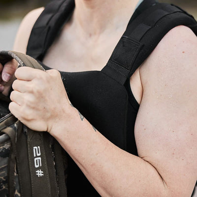 The individual, clad in a GORUCK Training Weight Vest 2.0 and holding a bag labeled 26#, seems to be outdoors, perhaps gearing up for an activity that demands physical strength. The image highlights their upper body and arms.