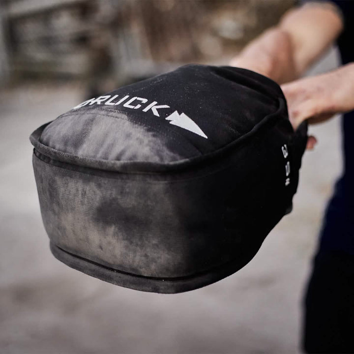 A person is holding a GORUCK Sand Kettlebell, featuring the brand name and arrow logo. The sand kettlebell displays signs of use, with visible wear marks that highlight its endurance design. The blurred background draws attention to this durable piece of equipment, similar in function to a portable kettlebell.