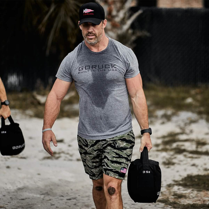 A man wearing a gray t-shirt and camo shorts strides along a sandy path outdoors, carrying two 35 lb GORUCK Sand Kettlebells. These black bags are ideal for endurance training. Behind him, a fence and trees can be seen. His shirt is noticeably damp with sweat.