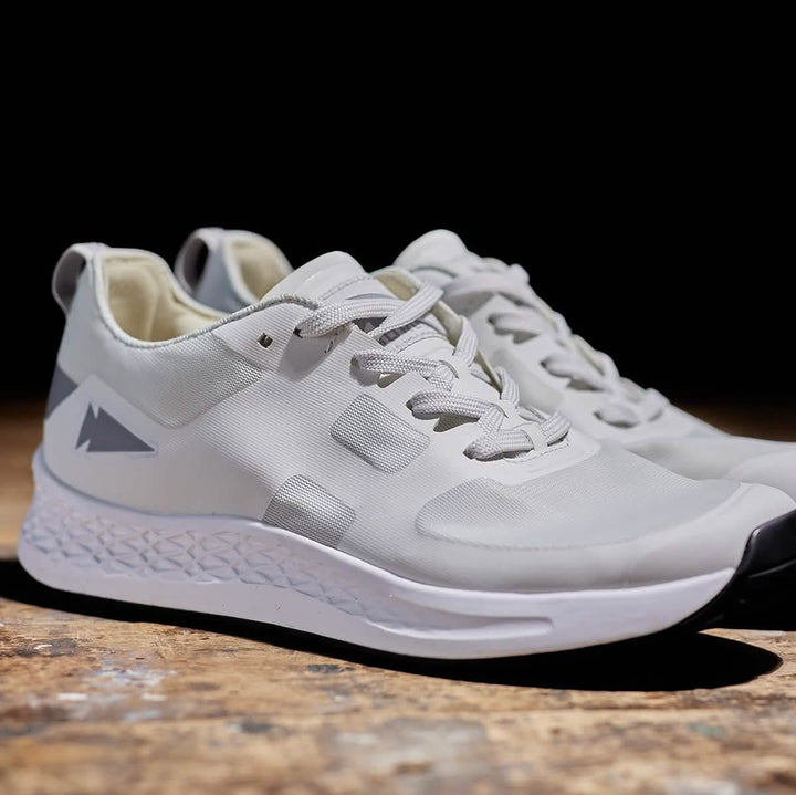 A pair of GORUCK Men's Rough Runner sneakers in light grey and dark grey, featuring a white gradient density sole and a triangular logo on the side, are placed on a wooden surface. The shoes include laces and a heel loop for easy wearing, with the dark background providing the perfect contrast to showcase these sneakers.