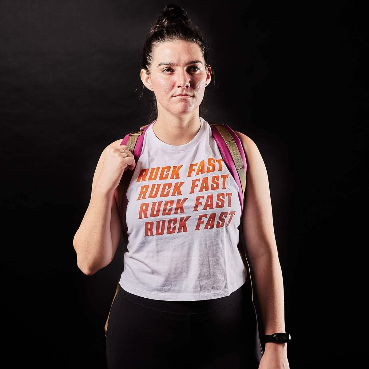 Against a dark background, an adventurous person with a bun hairstyle is confidently wearing the Women’s Ruck Fast Tank - Tri-Blend by vendor-unknown. The tank features bold orange "RUCK FAST" lettering and is complemented by a backpack strap casually worn over one shoulder.