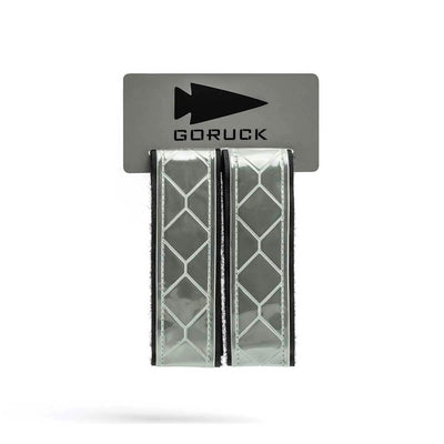 The GORUCK Reflective Ruck Bands, showcasing a diamond pattern, are affixed to a gray card emblazoned with the arrowhead logo and the brand name. Equipped with MOLLE webbing for secure attachment, these bands improve nighttime visibility, enhancing safety during outdoor activities.