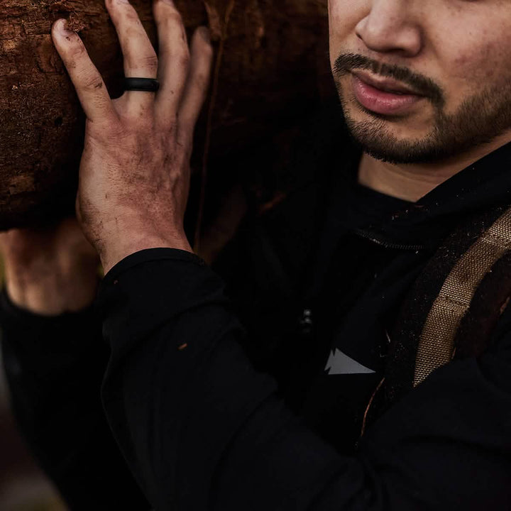 A man with dirty hands, dressed in dark clothing and a black ring, carries a large log on his shoulder while wearing the Indestructible Challenge Windbreaker made from abrasion-resistant ToughDry fabric.