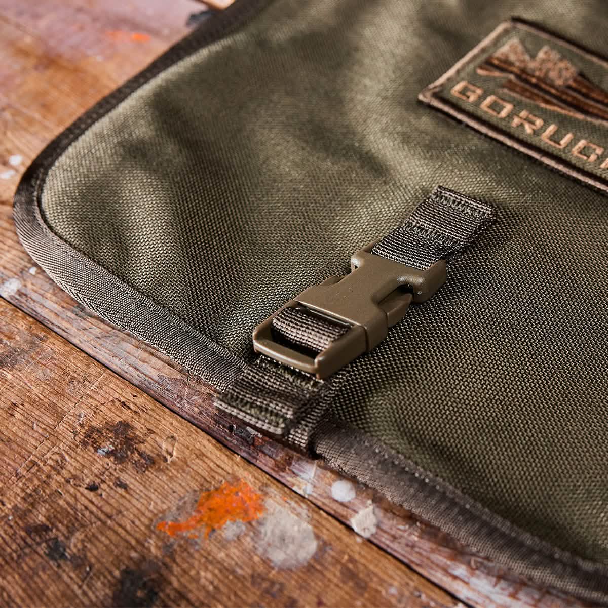 Close-up of the GORUCK Wire Dopp, showcasing its green fabric crafted from durable CORDURA. It features a plastic buckle and a patch on top, all resting on a paint-splattered wooden surface. Ideal for organized carry, the patch displays text and design.